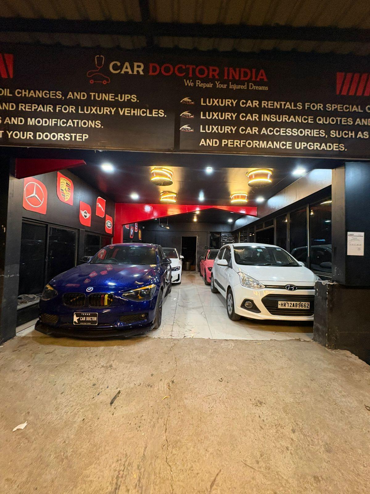 The Car Doctor India service bay at night with cars parked inside and luxury marque badges on the wall