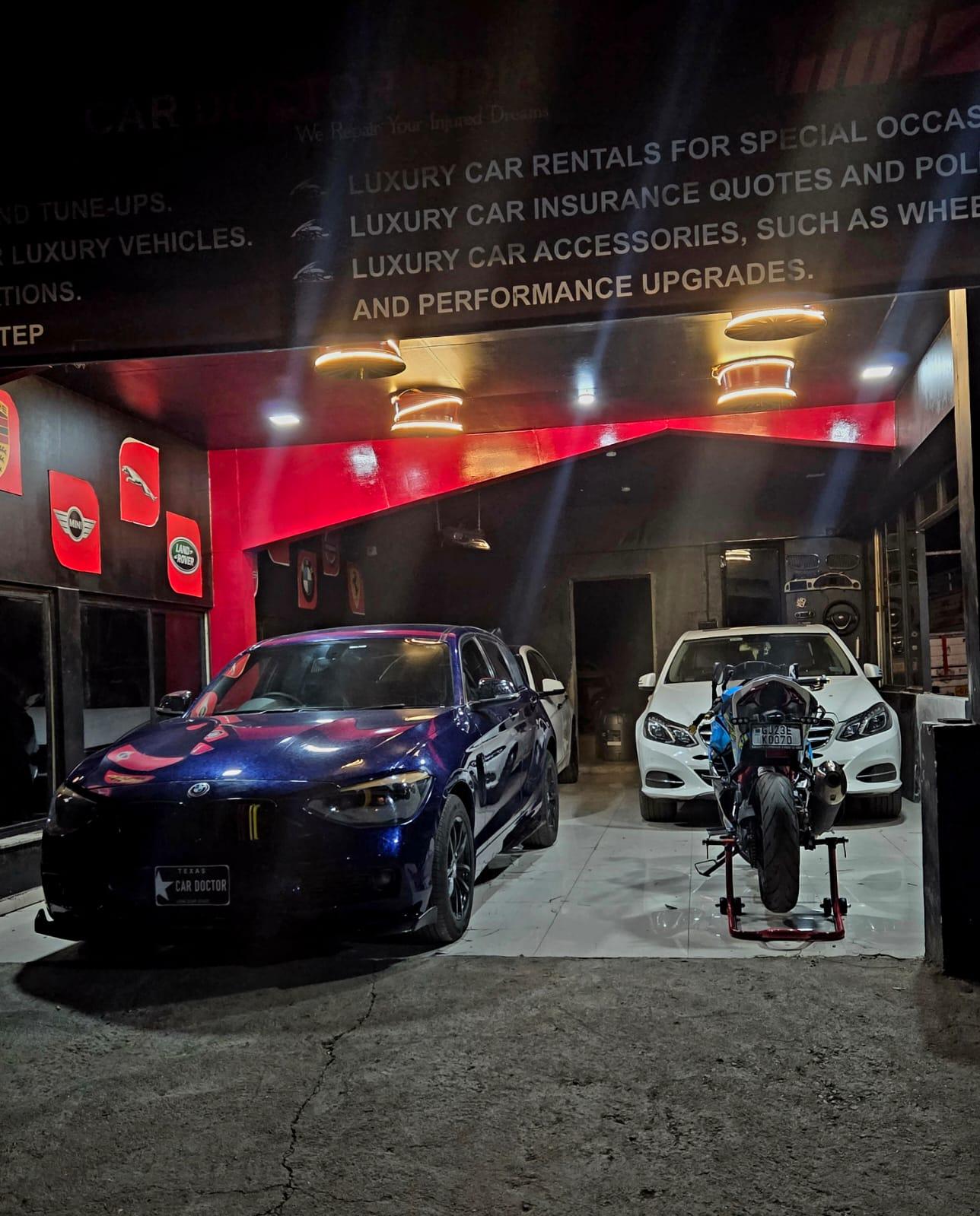 A dark blue BMW and a white Mercedes-Benz inside the Car Doctor India service bay at night, lit by red ambient lighting