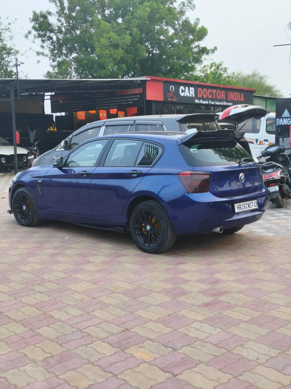 A blue BMW 1 Series with black alloy wheels and a rear spoiler parked outside the Car Doctor India workshop