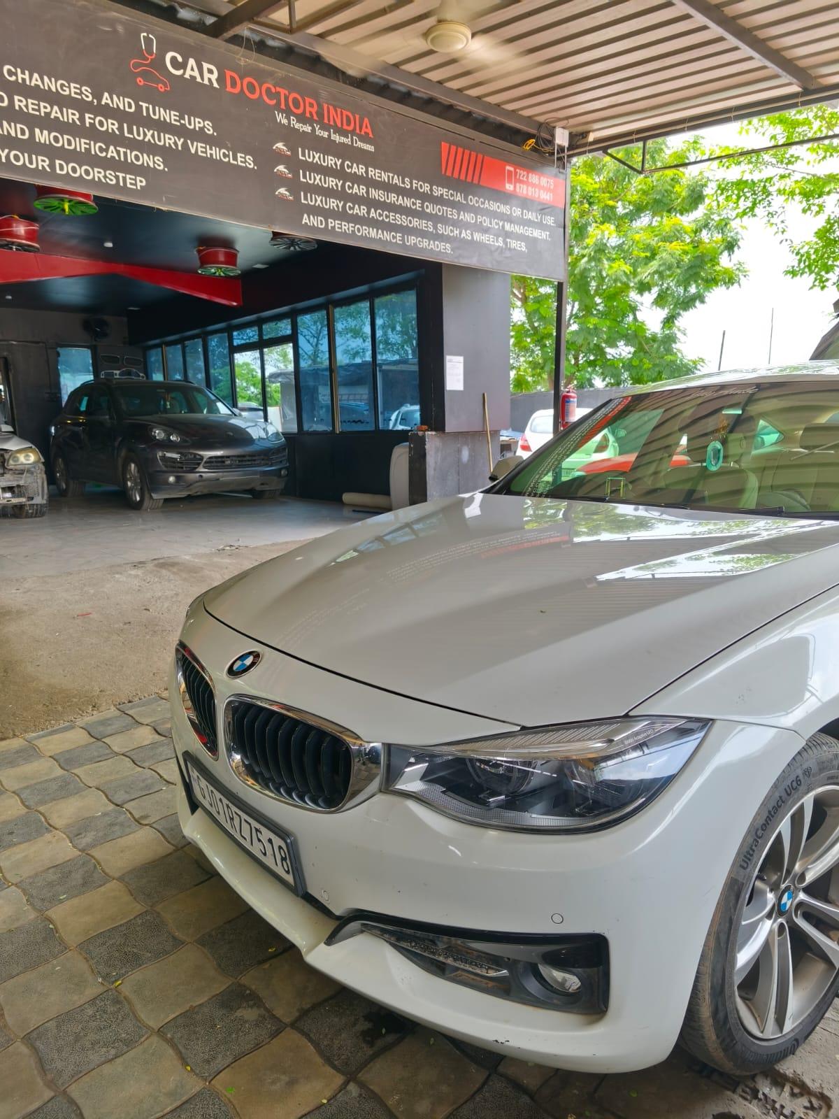 A white BMW 3 Series GT parked in front of the Car Doctor India workshop in daylight, with the service signage overhead and a Porsche Cayenne inside the bay behind