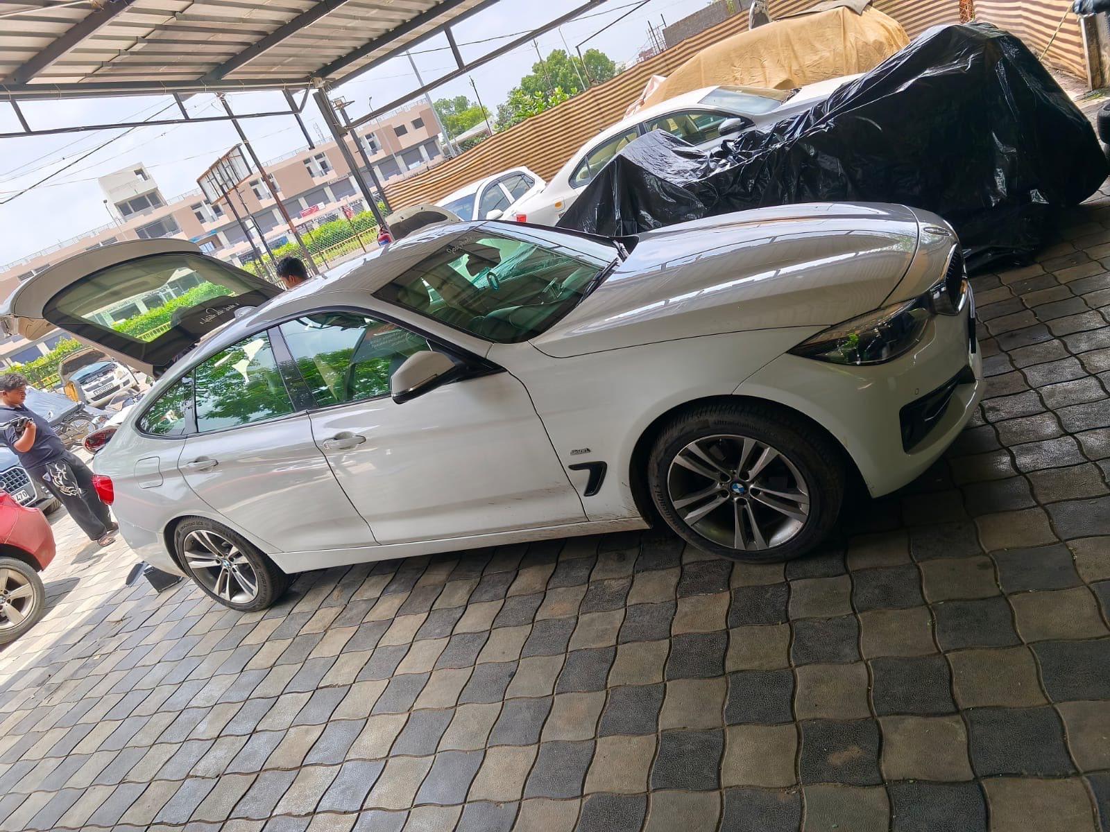 The side profile of a white BMW 3 Series GT parked under the Car Doctor India canopy, with covered cars waiting behind it