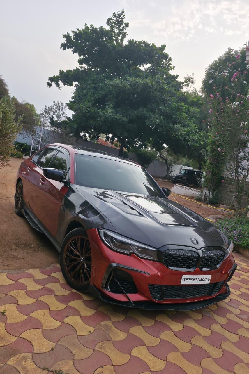 A red BMW M340i with an exposed carbon-fibre bonnet and front wings, a carbon front splitter and bronze alloy wheels, parked on paving beside flowering trees
